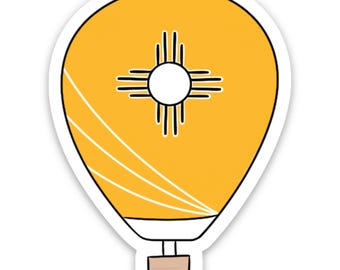 May include: A sticker featuring a yellow hot air balloon design. The balloon has a white outline and a New Mexico Zia symbol in the center. Three white curved lines are on the side, and a brown basket is at the bottom.