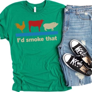 May include: Green t-shirt with a rooster, cow, and pig graphic and the text "I'd smoke that". The shirt is paired with blue jeans and black sneakers.
