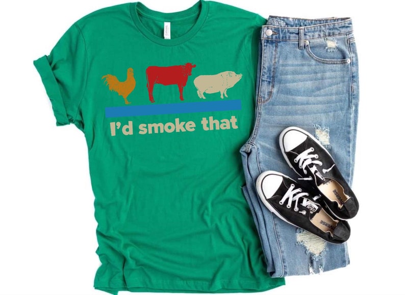 May include: Green t-shirt with a rooster, cow, and pig graphic and the text "I'd smoke that". The shirt is paired with blue jeans and black sneakers.