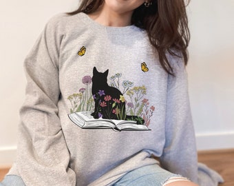 May include: A gray sweatshirt with a black cat silhouette sitting on an open book surrounded by colorful flowers. Two yellow butterflies are flying above the cat.