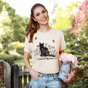 May include: A light peach-colored t-shirt featuring a black cat silhouette sitting amidst colorful flowers and butterflies, with an open book below. The design is printed on the front of the shirt. The shirt is short-sleeved.