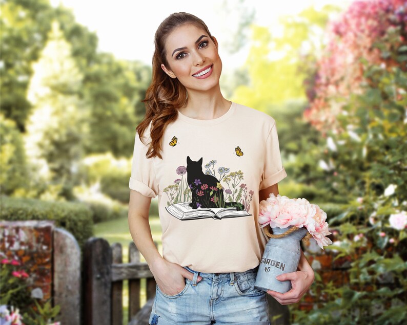 May include: A light peach-colored t-shirt featuring a black cat silhouette sitting amidst colorful flowers and butterflies, with an open book below. The design is printed on the front of the shirt. The shirt is short-sleeved.