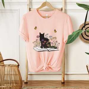 May include: A light pink t-shirt featuring a black cat silhouette sitting on an open book, surrounded by colorful flowers. Two yellow butterflies are flying near the cat. The shirt is hanging on a wooden rack.