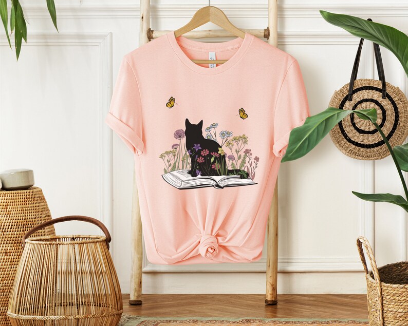 May include: A light pink t-shirt featuring a black cat silhouette sitting on an open book, surrounded by colorful flowers. Two yellow butterflies are flying near the cat. The shirt is hanging on a wooden rack.
