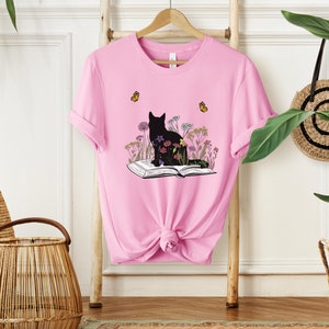May include: Pink t-shirt featuring a black cat silhouette sitting on an open book, surrounded by colorful flowers. Two yellow butterflies are flying above. The shirt is hanging on a wooden rack.