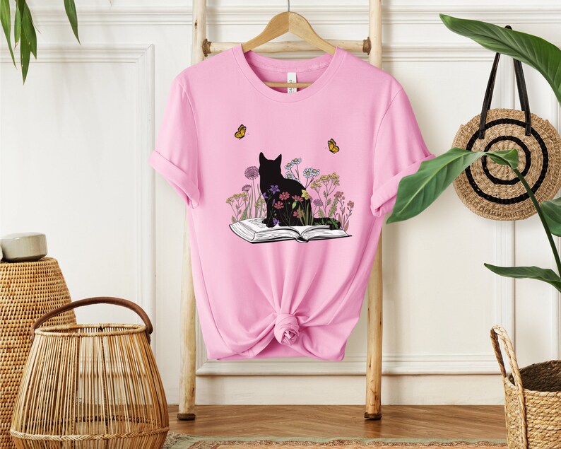 May include: Pink t-shirt featuring a black cat silhouette sitting on an open book, surrounded by colorful flowers. Two yellow butterflies are flying above. The shirt is hanging on a wooden rack.