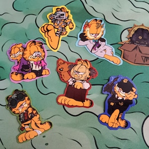 Peut inclure: Une collection d'autocollants Garfield colorés dans diverses poses et tenues. Les autocollants présentent le chat orange dans différents costumes, dont un robot, un policier et un personnage dans une boîte. Les autocollants sont sur une surface à motifs verts et bleus.