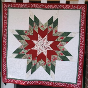 May include: A red and green quilted wall hanging featuring a large star design with a white background. The star is made up of multiple triangles and squares, and the fabric is patterned with cardinals, holly, and berries.