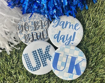 May include: Four circular pin-back buttons featuring blue and white designs. One button has a blue floral pattern with the text "GO BIG BLUE", another says "Game day!" in blue script, and two buttons feature the letters "UK" in blue patterns.