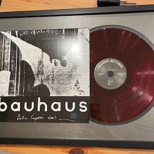 May include: A framed black and white photo of a band called Bauhaus with a red vinyl record in a silver frame. The record label is "Leaving Records" and the record is titled "In the Flat Field". The photo is signed "Bauhaus Bela Lugosi's Dead"