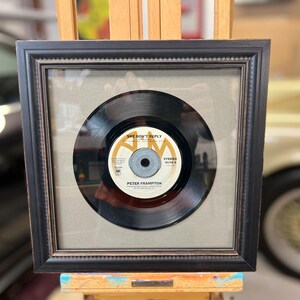 May include: A framed 45 RPM vinyl record by Peter Frampton. The record is black with a gold label. The label has the title "She Don't Reply" and the artist's name. The frame is black with a gold trim.