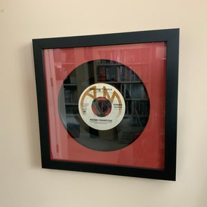 May include: Framed 45 RPM vinyl record of Peter Frampton's song "She Don't Reply." The record is displayed in a black frame with a red mat. The record label is white with gold and brown text. The frame is hanging on a beige wall.