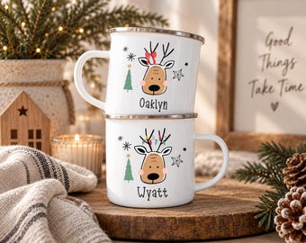 Könnte beinhalten: Zwei weiß emaillierte Tassen, die vertikal gestapelt sind und jeweils eine Rentier-Illustration mit einem grünen Tannenbaum, Sternen und Schneeflocken zeigen. Die obere Tasse trägt den Namen Oaklyn mit roter Schleife, die untere den Namen Wyatt mit bunter Lichterkette.