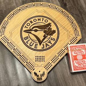 May include: A wooden baseball-shaped cribbage board featuring the Toronto Blue Jays logo, a deck of cards, and pegs. The board is light brown with engraved details and a series of holes for pegging. The deck of cards is red and white.