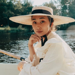 Op de afbeelding: Een vrouw met een brede strohoed met een zwart lint zit in een boot op een meer. Ze draagt een witte geplooide blouse en een witte rok.
