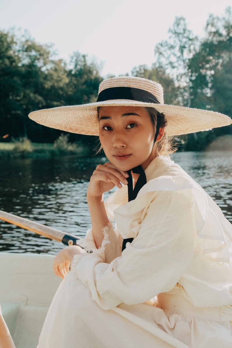 Op de afbeelding: Een vrouw met een brede strohoed met een zwart lint zit in een boot op een meer. Ze draagt een witte geplooide blouse en een witte rok.