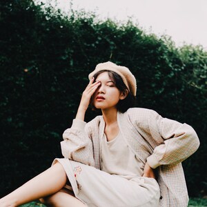 Op de afbeelding: Een vrouw met een beige baret, een geruit jasje en een witte rok met bloemenborduursel zit op een grasveld. Ze bedekt haar oog met haar hand.