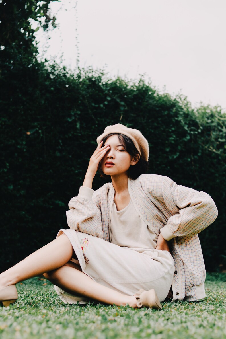 Op de afbeelding: Een vrouw met een beige baret, een geruit jasje en een witte rok met bloemenborduursel zit op een grasveld. Ze bedekt haar oog met haar hand.