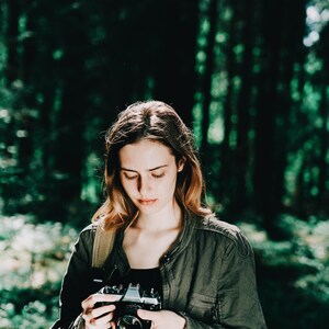 Op de afbeelding: Een vrouw in een groene jas houdt een vintage camera vast in een bosrijke omgeving. De camera is zwart en zilver met een lensdop.