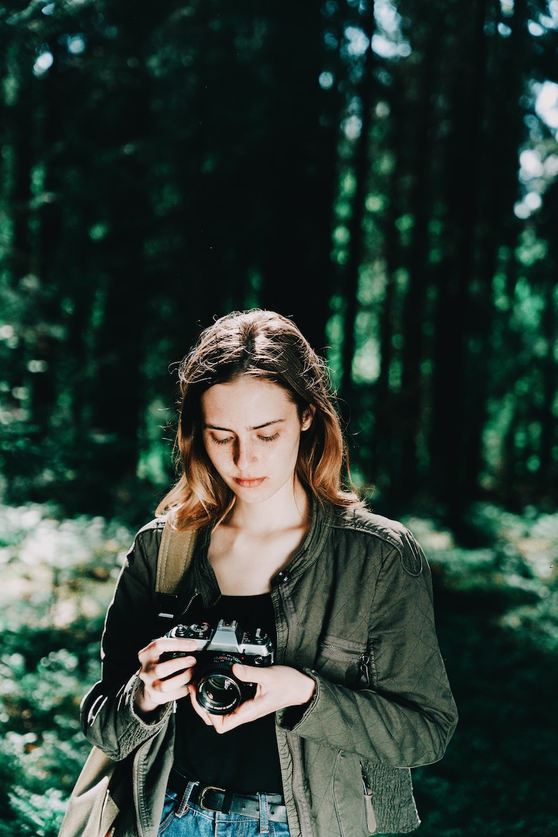 Op de afbeelding: Een vrouw in een groene jas houdt een vintage camera vast in een bosrijke omgeving. De camera is zwart en zilver met een lensdop.