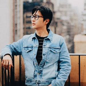 Op de afbeelding: Een man draagt een lichtblauw denim jack over een gestreept zwart-wit shirt, met blauwe jeans en een zwarte bril. Hij staat op een balkon met een houten balustrade.