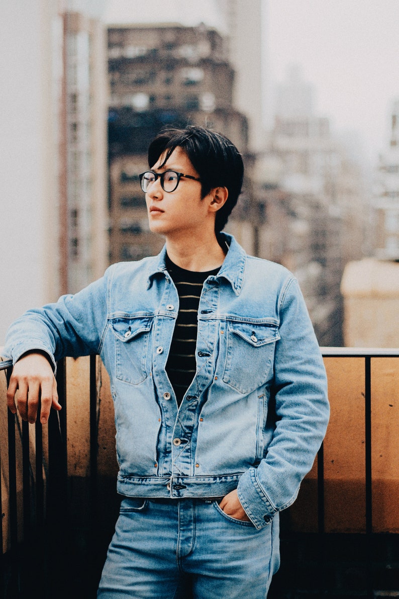 Op de afbeelding: Een man draagt een lichtblauw denim jack over een gestreept zwart-wit shirt, met blauwe jeans en een zwarte bril. Hij staat op een balkon met een houten balustrade.