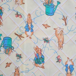 May include: A repeating pattern on a light yellow fabric featuring illustrations of rabbits, watering cans, birds, and floral elements. The rabbits are depicted in various poses, some wearing blue jackets. The design includes a grid pattern with light blue and brown accents.