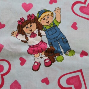 May include: Light blue fabric featuring a repeating pattern of pink hearts and cartoon children. The children are a girl in a pink skirt and a boy in blue overalls, each with a small brown dog. The fabric is likely for children's crafts.