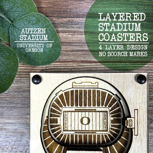May include: A wooden coaster featuring a layered design of Autzen Stadium, home of the University of Oregon Ducks. The coaster is made of four layers of wood and has a brown finish. The text "Autzen Stadium" and "University of Oregon" is printed on the bottom layer.