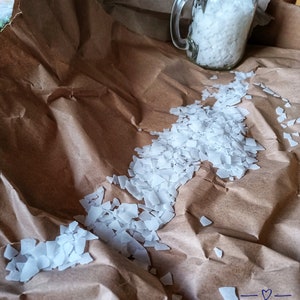 May include: A glass jar filled with white, irregularly shaped crystals sits on brown paper. The crystals are scattered around the jar, creating a trail.