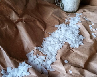 Può includere: Un barattolo di vetro pieno di cristalli bianchi di forma irregolare si trova su carta marrone. I cristalli sono sparsi intorno al barattolo, creando una scia.