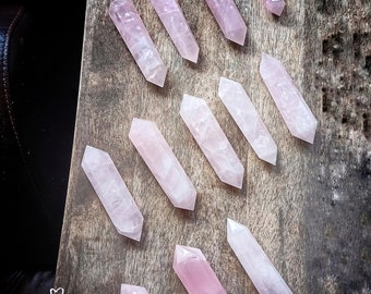 May include: A collection of 11 pink rose quartz crystal points arranged on a wooden surface. The points are all different sizes and shapes, and they are all polished to a smooth finish. The text "KASHMIR Luxuries" is visible in the lower left corner of the image.