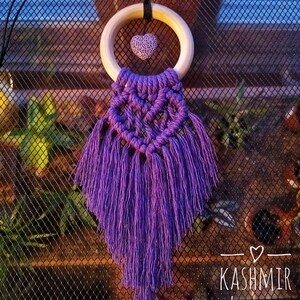 Op de afbeelding: Een paarse macramé wandhanger met een houten ring en een hartvormig accent. Het ontwerp heeft een geweven hartvorm en lange, vloeiende franjes. De tekst "Kashmir Luxuries" is zichtbaar.