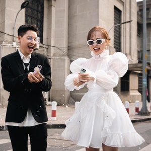 May include: A couple celebrates with confetti poppers outdoors. The person on the left wears a black suit with a white shirt and tie. The person on the right wears a white dress with puffy sleeves and sunglasses. The background features a city street and buildings.