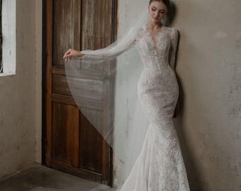 Può includere: Abito da sposa in pizzo bianco con maniche lunghe e silhouette a sirena. L'abito presenta uno scollo a V e dettagli di bottoni sul davanti. Un velo trasparente scende dalla testa e l'abito ha un lungo strascico.