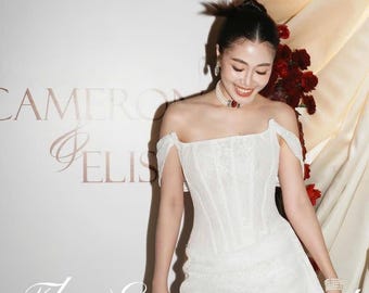 May include: A white, off-the-shoulder lace dress with a fitted bodice and a short skirt. The dress features a flowing train and is worn by a person. The words "The Engagement" are written in cursive on the bottom of the image.