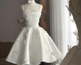 May include: A white, knee-length dress with butterfly appliqués, displayed on a mannequin. The dress features a fitted bodice, spaghetti straps, and a flared skirt. A gold crown and white bow are on the mannequin's head. The dress is likely for a special occasion.