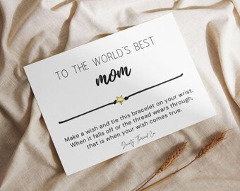 May include: A white card with the text "TO THE WORLD'S BEST mom" and a black string bracelet with a gold star charm. The card includes instructions and the brand name "Dainty Thread Co."