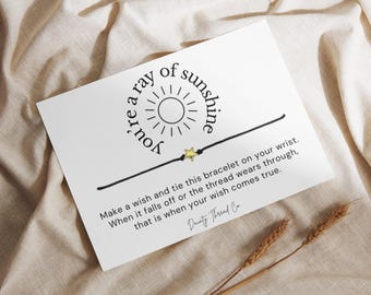 May include: A white card with the text "you're a ray of sunshine" and a black string bracelet with a gold star charm. The card includes instructions and is decorated with wheat stalks.
