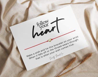 May include: A white card with the words "follow your heart" in black script. A red string bracelet with a gold heart charm is featured. The card includes text about making a wish. Two wheat stalks are in the foreground.