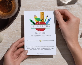 May include: A white card with a teacher appreciation design, held by hands. The design includes a red apple, pencils, a ruler, and the text "MS. MARTHA" and "THANK YOU FOR HELPING ME GROW." A black string bracelet with a gold star charm is attached.