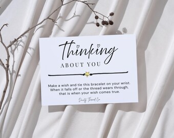 May include: A white card with the words "Thinking About You" and a black string bracelet with a gold star charm. The card includes instructions for making a wish. A sprig of a plant is in the background.