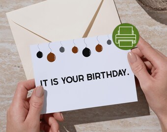 May include: A white birthday card with the text "IT IS YOUR BIRTHDAY." is held. The card has hanging baubles in black, brown, and grey. An envelope is in the background.