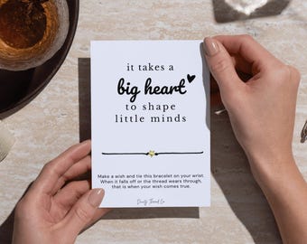 May include: A white card with the text "it takes a big heart to shape little minds" is held by two hands. A black string bracelet with a gold star charm is attached to the card. The card also includes instructions.