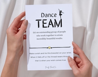 May include: A white card with the words "Dance TEAM" in black, a definition, and a wish bracelet. The bracelet has a black cord and a gold star charm. The card is held by hands.