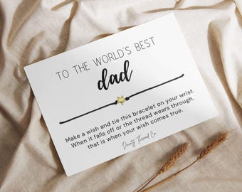 May include: A white card with the text "TO THE WORLD'S BEST dad" in black script. A black string bracelet with a gold star charm is featured. The card includes instructions and the brand name "Dainty Thread Co."