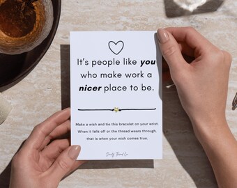 May include: A white card with a black heart and text that reads "It's people like you who make work a nicer place to be." A black string bracelet with a gold star charm is below the text. The card is held by two hands.