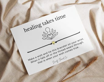 May include: A white card with the text "healing takes time" and a black string bracelet with a gold star charm. The card includes instructions and the brand name "Dainty Thread Co."