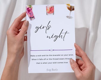 May include: A white card with the words "girls night" in cursive script, featuring watercolor illustrations of cocktails. A purple string bracelet with a star charm is attached. The card includes instructions for tying the bracelet.