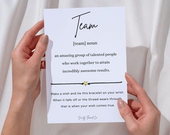 May include: A white card with the word "Team" in script font, with a definition below. A black string bracelet with a gold star charm is attached to the card. The card is held by hands.